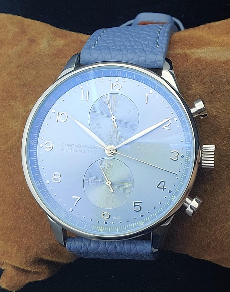 Puede incluir: Un reloj cron&oacute;grafo plateado con esfera azul claro y correa a juego. El reloj tiene una esfera redonda con manecillas y n&uacute;meros plateados, y dos esferas m&aacute;s peque&ntilde;as. El texto "CHRONOGRAPH AUTOMATIC" es visible.