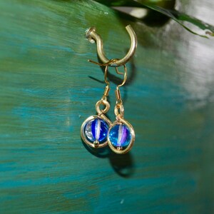 May include: A pair of dangle earrings featuring gold-toned metal hooks and circular frames. Each earring contains a spherical glass bead with a translucent blue and white striped pattern, suspended within the center of the gold-toned circular wire frame.