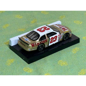 Op de afbeelding: Een goud-wit model van een raceauto met nummer 23 op de zijkant en het dak. De auto heeft sponsorlogo's en de woorden "Winston Lights" en "Food City". Het model staat op een zwarte basis.