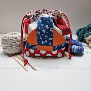 May include: A drawstring project bag constructed from patchwork fabric featuring a red base with white polka dots, orange panels, and a blue section with a white leaf pattern. The top is finished with a floral print fabric and red cords.