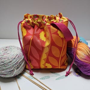 May include: A fabric drawstring bag featuring a vibrant orange and red floral pattern with yellow accents. The bag has a yellow interior lining with a small red flower print, a pink woven shoulder strap, and two pink cords for closure.