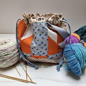 May include: A drawstring project bag featuring a patchwork design with orange, white, and light blue floral fabric panels. The base has a tan and black plaid pattern, and the top closure is a cream fabric with a dark brown floral print. It includes blue cord drawstrings and a matching blue shoulder strap.