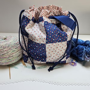 May include: A drawstring project bag featuring a patchwork design of blue and white floral fabric squares with quilted stitching. The bag has a blue fabric lining, a blue woven handle, and dark blue cord drawstrings with knotted ends.