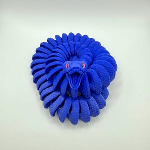 Pode incluir: Escultura de cobra azul enrolada, com um corpo texturizado e detalhado. A cabe&ccedil;a da cobra est&aacute; levantada, revelando uma boca bem aberta e olhos rosa. A escultura est&aacute; sobre um fundo branco liso.