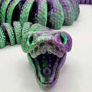 Pode incluir: Um brinquedo de cobra articulada impresso em 3D em tons de verde e roxo. A cabe&ccedil;a da cobra est&aacute; aberta, revelando um interior detalhado. Os olhos s&atilde;o verde brilhante. O corpo &eacute; segmentado, permitindo um movimento flex&iacute;vel. Um brinquedo &uacute;nico e colorido.