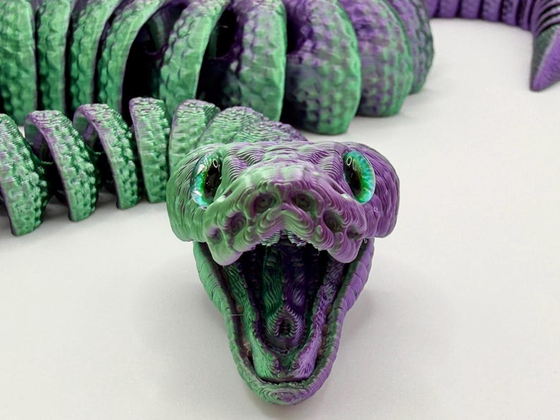 Pode incluir: Um brinquedo de cobra articulada impresso em 3D em tons de verde e roxo. A cabe&ccedil;a da cobra est&aacute; aberta, revelando um interior detalhado. Os olhos s&atilde;o verde brilhante. O corpo &eacute; segmentado, permitindo um movimento flex&iacute;vel. Um brinquedo &uacute;nico e colorido.