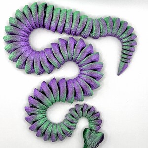 Pode incluir: Uma escultura decorativa de cobra em tons de roxo e verde. O corpo segmentado curva-se num padr&atilde;o sinuoso, com cada segmento exibindo uma superf&iacute;cie texturizada. A cabe&ccedil;a &eacute; detalhada com um padr&atilde;o de escamas, e o design geral &eacute; art&iacute;stico.