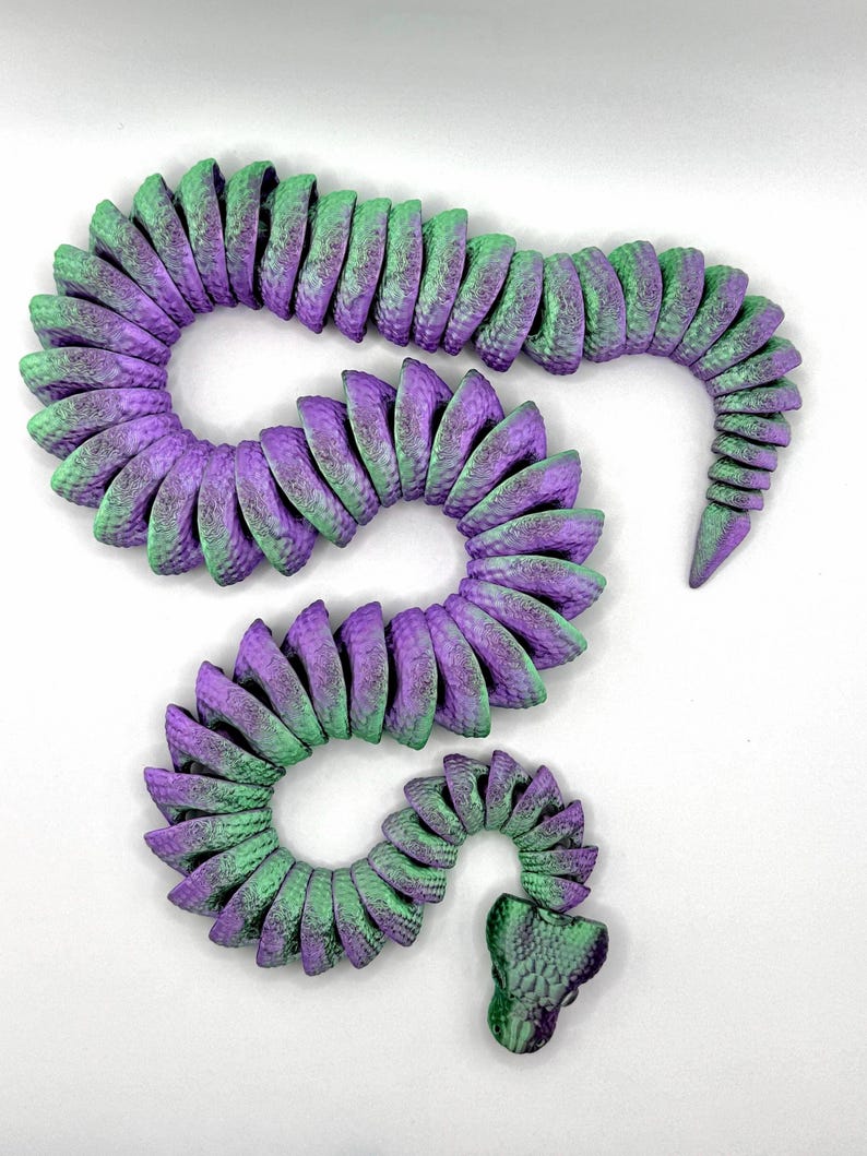 Pode incluir: Uma escultura decorativa de cobra em tons de roxo e verde. O corpo segmentado curva-se num padr&atilde;o sinuoso, com cada segmento exibindo uma superf&iacute;cie texturizada. A cabe&ccedil;a &eacute; detalhada com um padr&atilde;o de escamas, e o design geral &eacute; art&iacute;stico.