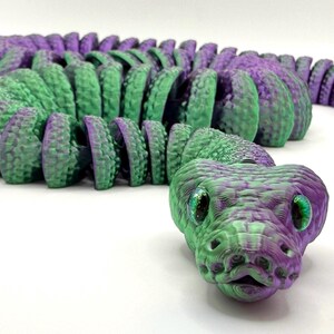 Pode incluir: Um brinquedo de cobra articulada impresso em 3D em tons de verde e roxo. A cabe&ccedil;a da cobra est&aacute; em foco, com escamas detalhadas e olhos verdes brilhantes. O corpo &eacute; segmentado, permitindo um movimento flex&iacute;vel. O brinquedo foi projetado para se assemelhar a uma cobra realista.