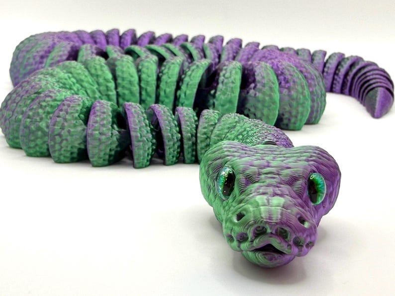 Pode incluir: Um brinquedo de cobra articulada impresso em 3D em tons de verde e roxo. A cabe&ccedil;a da cobra est&aacute; em foco, com escamas detalhadas e olhos verdes brilhantes. O corpo &eacute; segmentado, permitindo um movimento flex&iacute;vel. O brinquedo foi projetado para se assemelhar a uma cobra realista.