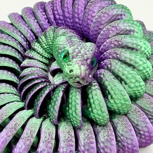 Pode incluir: Escultura de cobra impressa em 3D em tons de roxo e verde. O corpo da cobra &eacute; enrolado, com um padr&atilde;o de escamas detalhado. A cobra tem olhos verdes e uma superf&iacute;cie texturizada, criando uma pe&ccedil;a decorativa &uacute;nica.