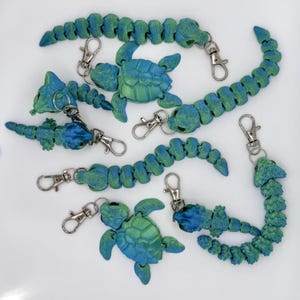 Op de afbeelding: Een verzameling blauwgroene sleutelhangers in de vorm van schildpadden en beweeglijke slangen, met zilverkleurige sluitingen. De sleutelhangers zijn gemaakt van plastic en hebben een textuuroppervlak. De schildpadden hebben een schildontwerp.