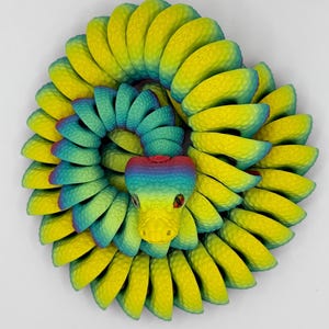 Pode incluir: Escultura de cobra estilizada e colorida, em forma espiral. Apresenta um gradiente de cores amarelo, verde e azul. A cabe&ccedil;a da cobra &eacute; amarela com olhos vermelhos. Uma pe&ccedil;a de arte &uacute;nica.