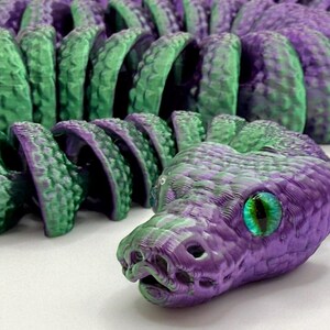 Pode incluir: Um brinquedo de cobra articulada impresso em 3D em tons de roxo e verde. A cabe&ccedil;a da cobra apresenta um design detalhado com a boca aberta e um grande olho verde e azul iridescente. O corpo &eacute; segmentado, permitindo um movimento flex&iacute;vel.