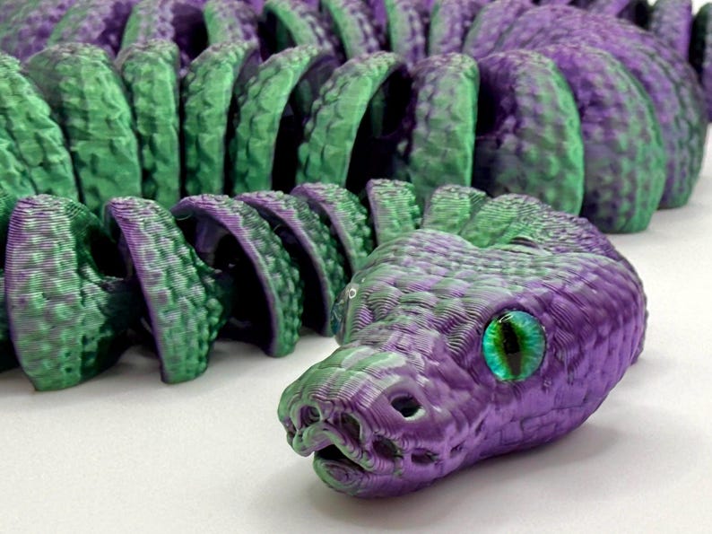 Pode incluir: Um brinquedo de cobra articulada impresso em 3D em tons de roxo e verde. A cabe&ccedil;a da cobra apresenta um design detalhado com a boca aberta e um grande olho verde e azul iridescente. O corpo &eacute; segmentado, permitindo um movimento flex&iacute;vel.
