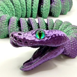 Pode incluir: Um brinquedo de cobra articulada impresso em 3D em tons de roxo e verde. A cabe&ccedil;a da cobra &eacute; roxa com uma textura detalhada e um olho verde e preto. O corpo alterna entre segmentos roxos e verdes.