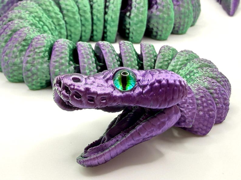 Pode incluir: Um brinquedo de cobra articulada impresso em 3D em tons de roxo e verde. A cabe&ccedil;a da cobra &eacute; roxa com uma textura detalhada e um olho verde e preto. O corpo alterna entre segmentos roxos e verdes.