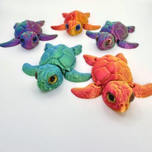 May include: Six colorful 3D-printed sea turtles in shades of purple, orange, green, and red. Each turtle has detailed shell patterns and large, expressive eyes. The turtles are arranged on a white surface.