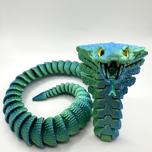 May include: A segmented, articulated snake toy in shades of blue and green. The head is a vibrant blue with yellow eyes and an open mouth revealing a forked tongue. The body coils and curves, showcasing a gradient of colors.