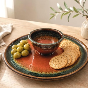 May include: A round ceramic serving platter with a deep orange center and a dark green rim, featuring a matching small bowl in the center. The platter holds a cluster of green olives and a stack of round, seeded crackers on its glazed surface.