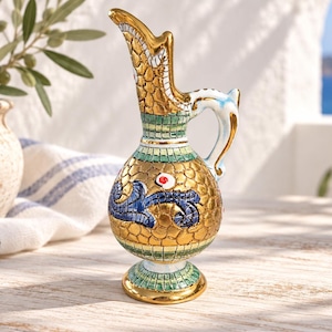 May include: A ceramic pitcher featuring a gold-toned, textured surface with a mosaic-like pattern. The pitcher includes green and white accents, a blue decorative swirl, a small red dot detail, and a curved handle with a gold-trimmed rim.