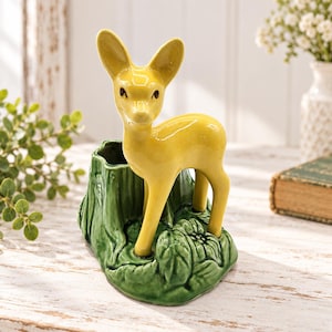 May include: A yellow ceramic deer figurine standing on a green base shaped like a tree stump with sculpted leaves. The deer has large ears and dark eyes, and the base features a hollow opening, functioning as a small planter or holder.