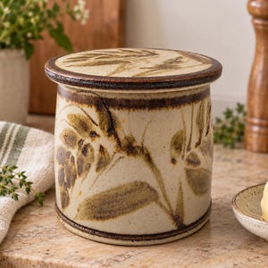 May include: A cylindrical ceramic butter crock with a lid, featuring a speckled cream-colored glaze and hand-painted brown botanical leaf motifs. The rim and lid edge are finished with a dark brown band, creating a rustic appearance for this kitchen vessel.