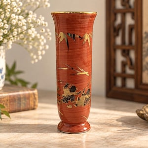 May include: A tall, cylindrical vase with a reddish-brown wood grain finish, featuring a gold-rimmed top edge. The exterior is decorated with gold-colored bamboo leaf motifs and a detailed scene of figures in black and gold near the base.