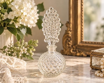 Puede incluir: Un frasco de perfume de vidrio transparente con una base esférica que presenta una textura de puntos en relieve. El frasco incluye un tapón alto y puntiagudo adornado con un patrón de puntos a juego, creando un diseño coherente en el vidrio.