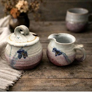 May include: A ceramic sugar bowl with a lid and a matching creamer pitcher. Both pieces feature a speckled off-white glaze with a gradient of dusty pink at the base and a hand-painted blue floral motif. The sugar bowl has a curved handle on the lid.