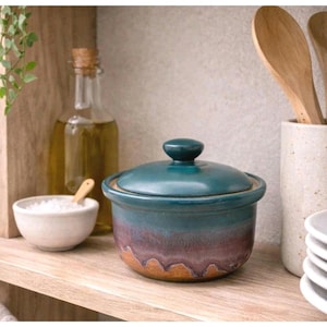 May include: A round ceramic lidded jar featuring a gradient glaze that transitions from a deep teal at the top to a muted purple and earthy brown at the base. The lid has a small knob handle, and the jar sits on a wooden shelf.