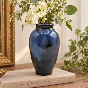 May include: A glossy, deep blue ceramic vase with a smooth, rounded body and a narrow neck. The vase features a gradient color effect, transitioning from a dark navy base to a lighter, mottled blue near the top, and is filled with white flowers.