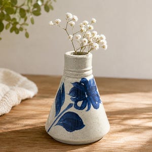 May include: A conical ceramic vase with a speckled off-white finish, featuring a hand-painted dark blue floral design with leaves and petals. The vase has a narrow neck with a subtle ridge and contains a small sprig of white baby's breath flowers.