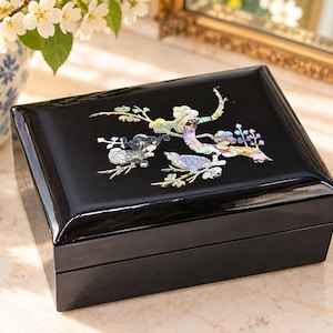 May include: A rectangular black lacquer jewelry box featuring an iridescent mother-of-pearl inlay on the lid. The inlay depicts two birds perched on flowering branches with shimmering shades of purple, green, and blue against the dark surface.
