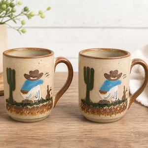 May include: Two ceramic mugs with brown handles and a beige speckled finish. Each mug features a cartoon cowboy design with a cactus and a brown and green landscape. The mugs are suitable for coffee or tea.