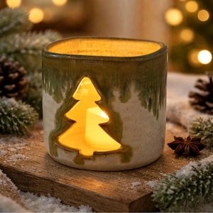May include: A cylindrical ceramic candle holder with a cream-colored base and a green glazed top. A cutout in the shape of a pine tree is centered on the front, revealing a glowing yellow candle flame inside the vessel, creating a warm light.