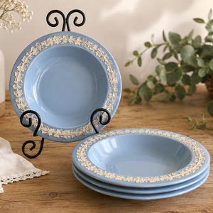 May include: A set of light blue ceramic bowls featuring a raised white floral vine pattern along the rim. One bowl is displayed upright on a black metal stand, while three additional bowls are stacked horizontally on a wooden surface.
