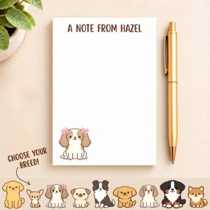 May include: A white rectangular notepad with the brown text A NOTE FROM HAZEL at the top and a cartoon illustration of a brown and white dog with pink bows in the bottom left corner, accompanied by a gold metal pen placed to the right side.