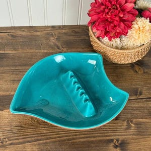 May include: A teal ceramic ashtray with a leaf-like shape and a central raised ridge featuring five notches for resting items. The glossy finish reflects light, highlighting the smooth, curved edges of the bowl-shaped design.