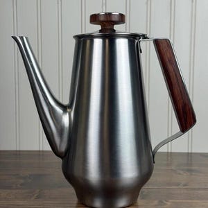 May include: A stainless steel coffee pot with a brushed metal finish, featuring a long, tapered spout and a dark brown wood-grain handle. The lid is topped with a matching circular wood-grain knob, providing a contrast to the metallic body.