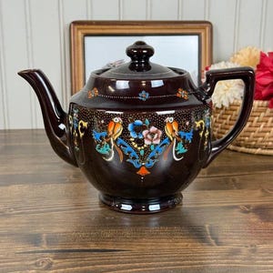 May include: A dark brown ceramic teapot featuring a glossy finish and a hand-painted design of colorful birds, blue floral scrolls, and small orange accents. The teapot includes a matching lid, a curved spout, and a sturdy handle on the side.