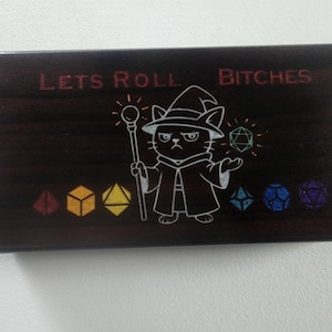 May include: A rectangular wooden box with the text "LETS ROLL BITCHES" in red. It features a cartoon cat wizard with a staff and dice illustrations in various colors, perfect for tabletop gaming enthusiasts.
