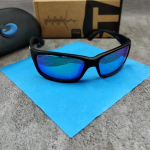 May include: Black sunglasses with blue mirrored lenses rest on a blue square cloth. The sunglasses have a thick frame and curved temples. A black case with a blue logo and a brown cardboard box are in the background.