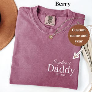 May include: Berry-colored t-shirt with the text "Sophia's Daddy EST. 2026" in white. The shirt has a crew neck and a Comfort Colors tag. A gold necklace with a pendant is draped across the shirt.