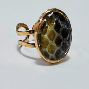 May include: A gold-toned ring featuring an oval-shaped setting with a textured, multi-colored inlay that transitions from olive green to dark brown in a scale-like pattern. The band is open and adjustable, constructed from a thin, polished metal wire.
