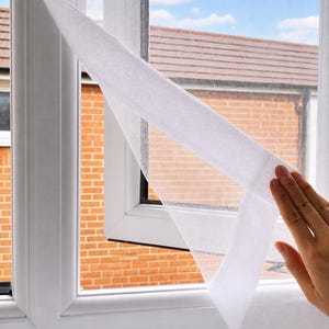 May include: A white mesh window screen with a thick adhesive border attached to a white window frame. The fine, semi-transparent netting is partially peeled back from the frame, revealing the grid-like texture of the material and the window.