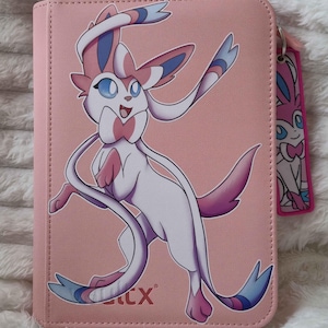 Puede incluir: Estuche rosa con una ilustración del personaje Sylveon. El estuche tiene un cierre de cremallera y una etiqueta a juego con un diseño Sylveon más pequeño. El personaje Sylveon es blanco, rosa y azul.