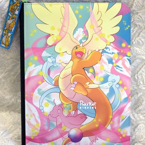 May include: A rectangular trading card binder featuring a large orange dragon-like creature with wings and a tail orb. The background displays pink ribbons, blue clouds, and yellow stars. The text PokeKat BINDERS is printed in white. A matching zipper pull is attached.