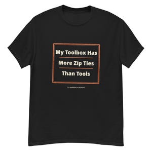 May include: A black short-sleeve t-shirt featuring a rectangular orange-bordered graphic on the chest. The text inside reads: My Toolbox Has More Zip Ties Than Tools. Below the graphic, the text LA BARRANCA DESIGNS is printed in small white letters.