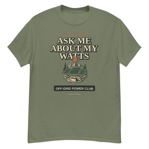 Op de afbeelding: Olijfgroen t-shirt met de tekst "ASK ME ABOUT MY WATTS" boven een illustratie van zonnepanelen, een berg en een zon. Onder de illustratie staat de tekst "OFF-GRID POWER CLUB".